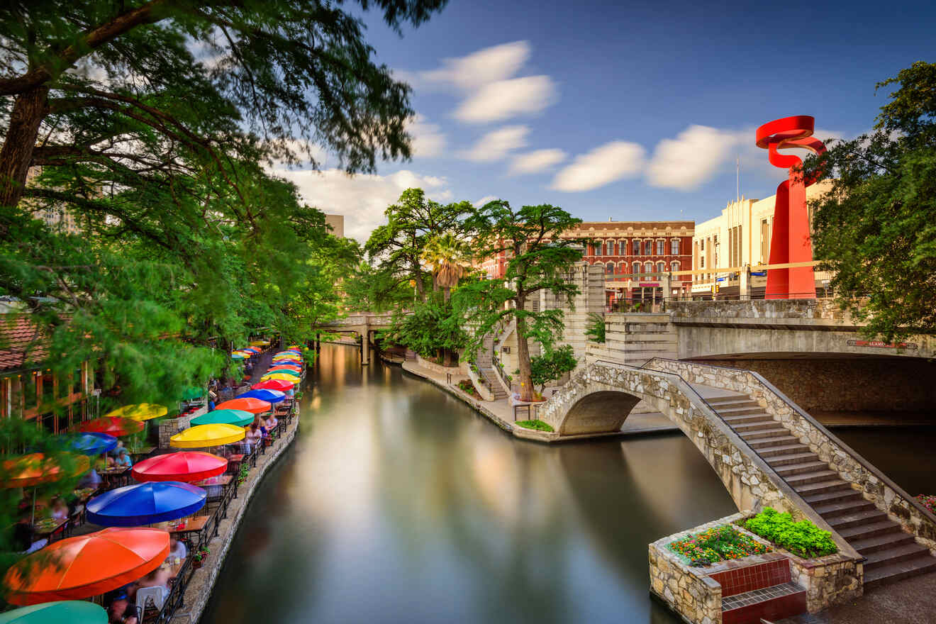 Long-exposure shot of San Antonio Riverwalk with vibrant umbrellas lining the bank and a striking red sculpture above the bridge